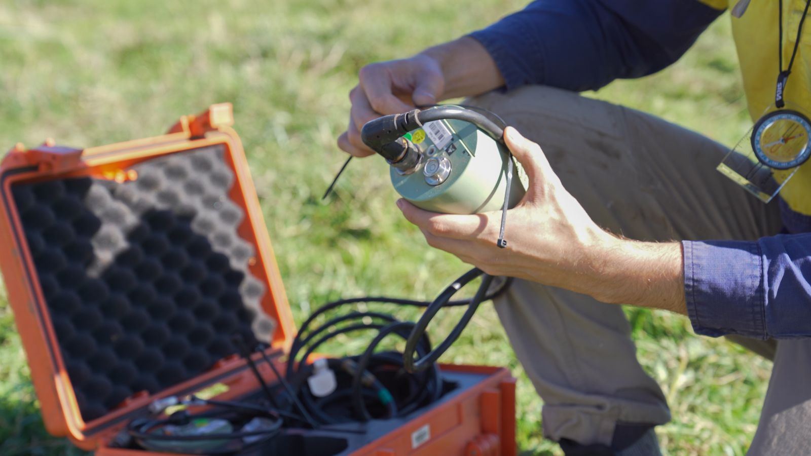 A technician crouches over a storage box in a grassy field, preparing a seismic sensor for insertion.